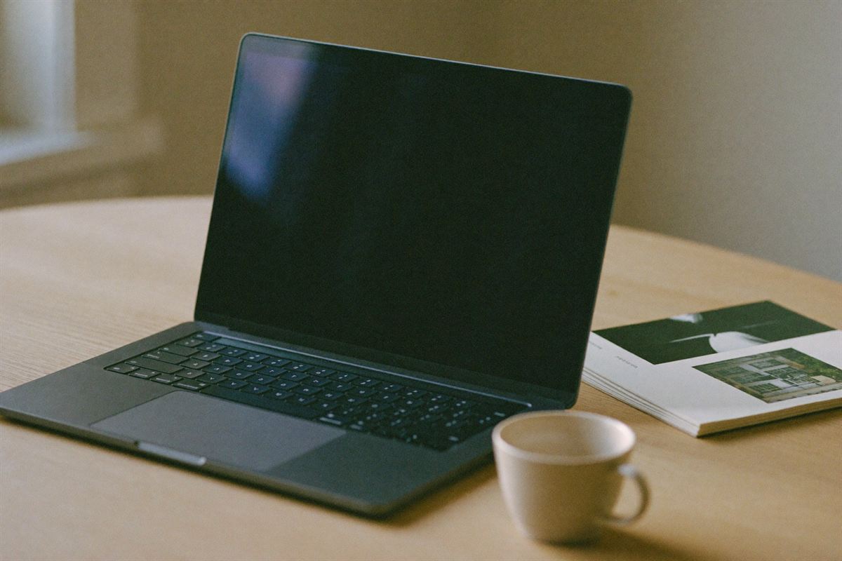 An open laptop on a pale oak desk beside a small stack of printed photographs and a ceramic cup, in soft window light.