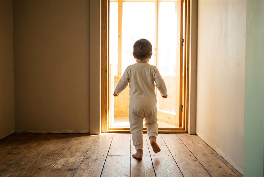 A young child running barefoot across wooden floorboards toward a bright open doorway, seen from behind.