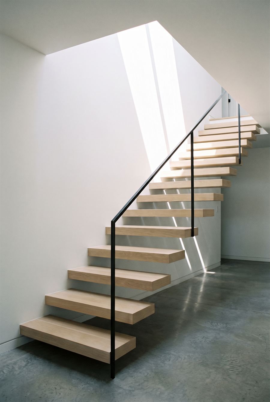 An open-tread pale oak staircase against a white wall, lit from a skylight above.