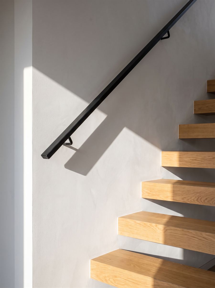 A pale oak open-tread staircase meeting a white plaster wall, with a slim black handrail and raking sunlight.